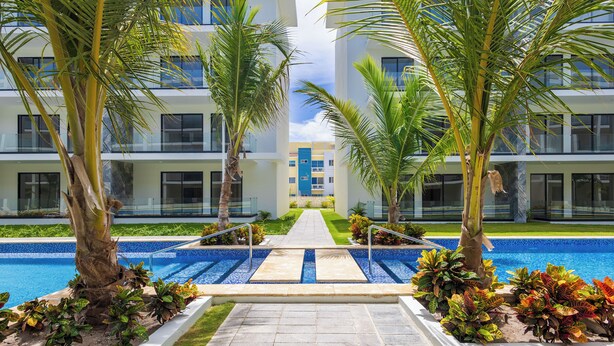 A modern hotel with a central pool, surrounded by palm trees and a well-maintained garden. - Tropical 2BR Retreat - Terrace, BBQ and Pool (Punta Cana)