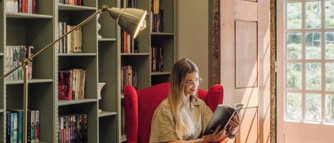 Una persona sentada en una silla roja frente a una pequeña mesa, con un libro y un juego de mesa, en una habitación con una estantería y un ventanal grande.