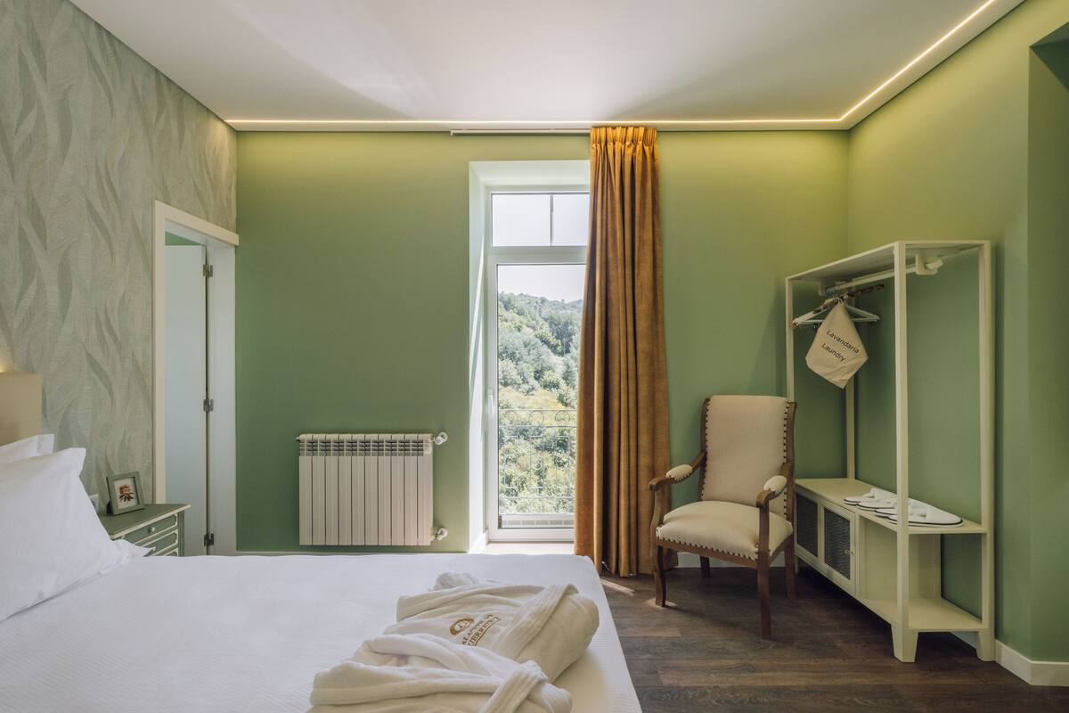 A hotel room with a bed, a chair, a robe rack, and a window with curtains.