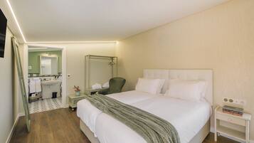 Hotelkamer met een groot bed, nachtkastje met een boek, een stoel, een spiegel en een badkamer door een geopende deur zichtbaar.