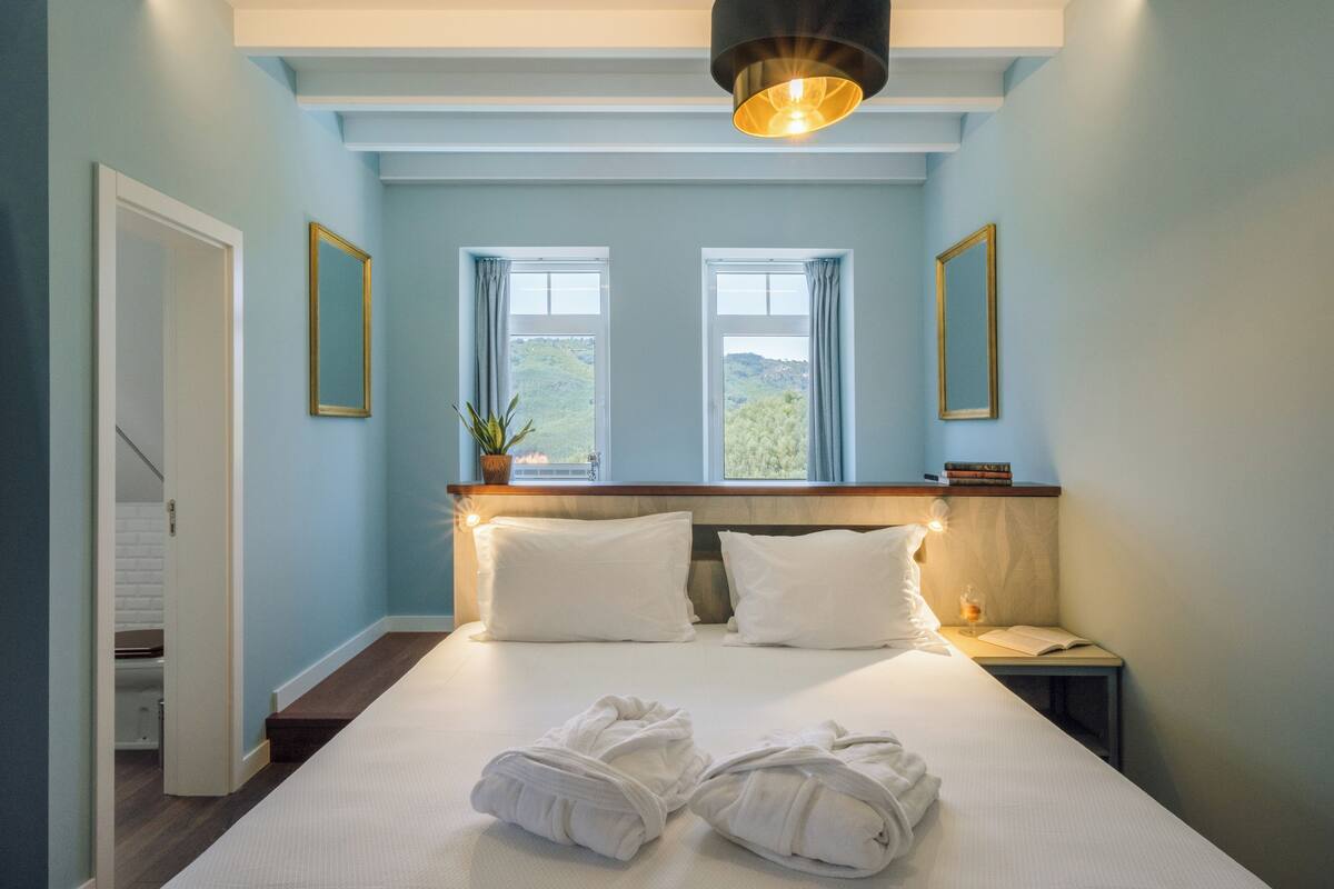 A bedroom with a bed, two windows, a nightstand, and a potted plant.