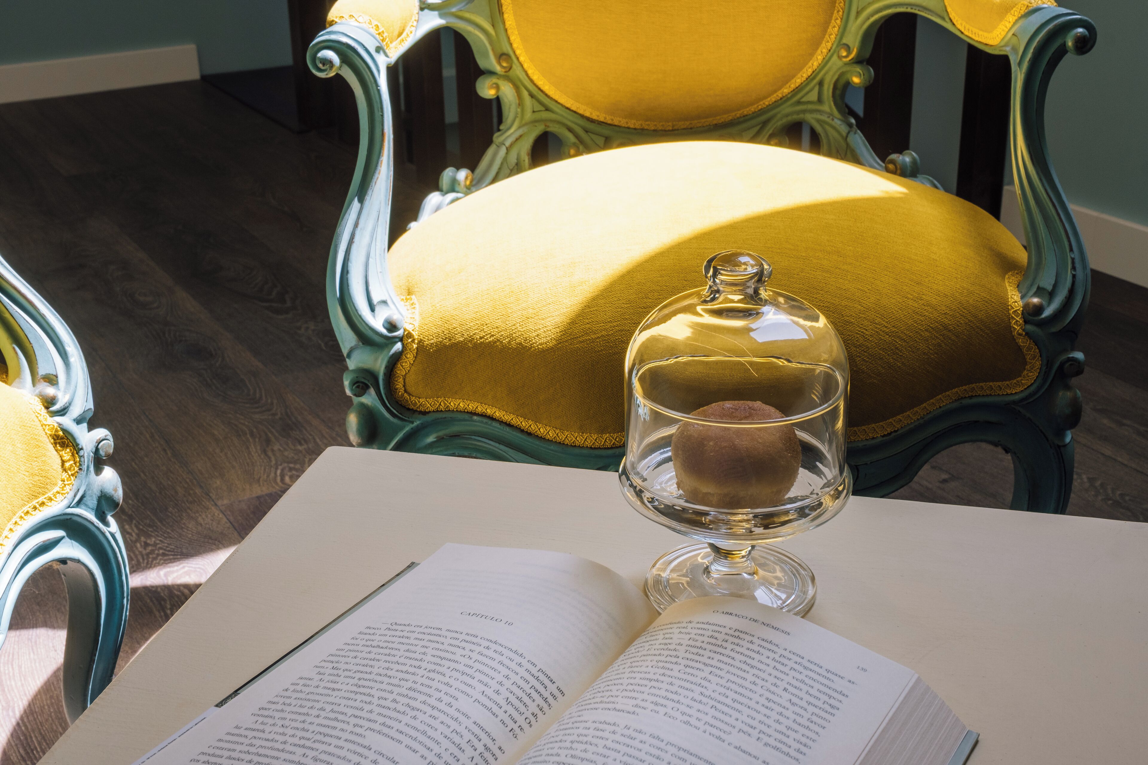 An open book on a table with a glass dome over a candle. A chair with a yellow cushion is in the background.