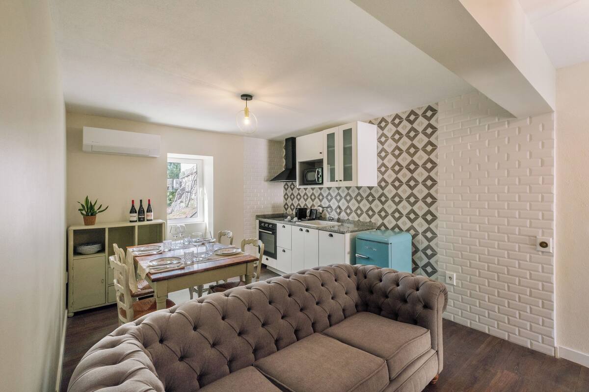 A compact kitchen with white cabinets, a microwave, and a sink. There is a dining area with a table set for four, a tufted sofa, and a potted plant.
