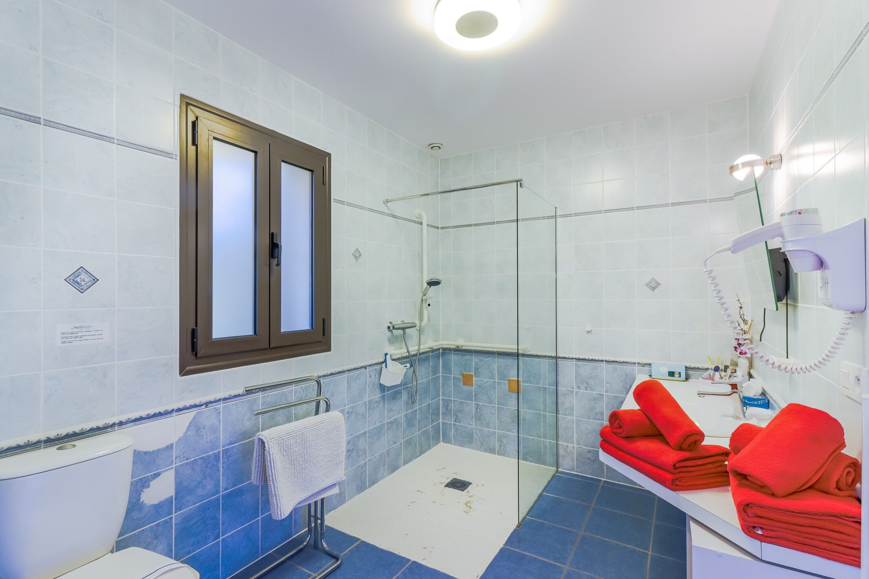 A bathroom with a glass shower enclosure, a toilet, a sink with red towels, and a mirror with a telephone.