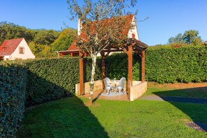 Ein Kiosk mit Tisch und Stühlen, umgeben von einer gepflegten Hecke und Rasenfläche.