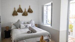 Un llit ben fet amb llençols grisos i almoades, una petita taula auxiliar de fusta i una cadira de vímet.
