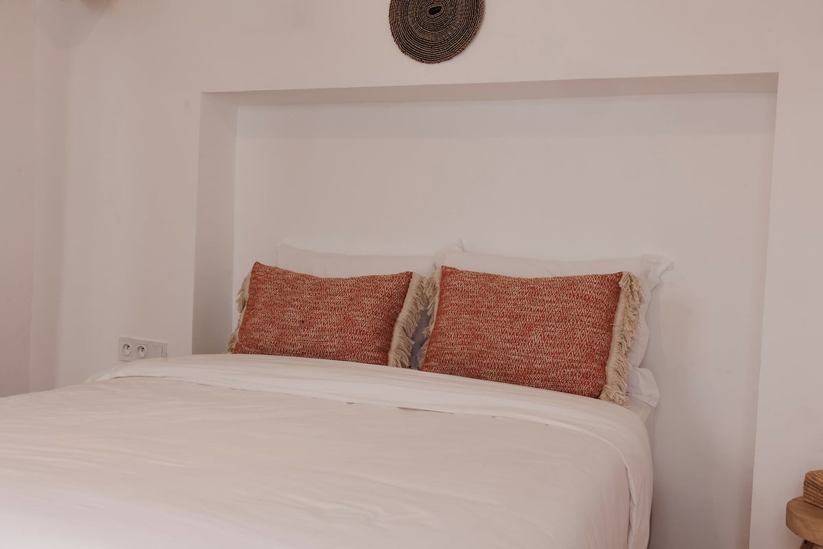 A bed with white bedding and two patterned pillows, a power outlet on the wall, and a woven wall hanging above the bed.