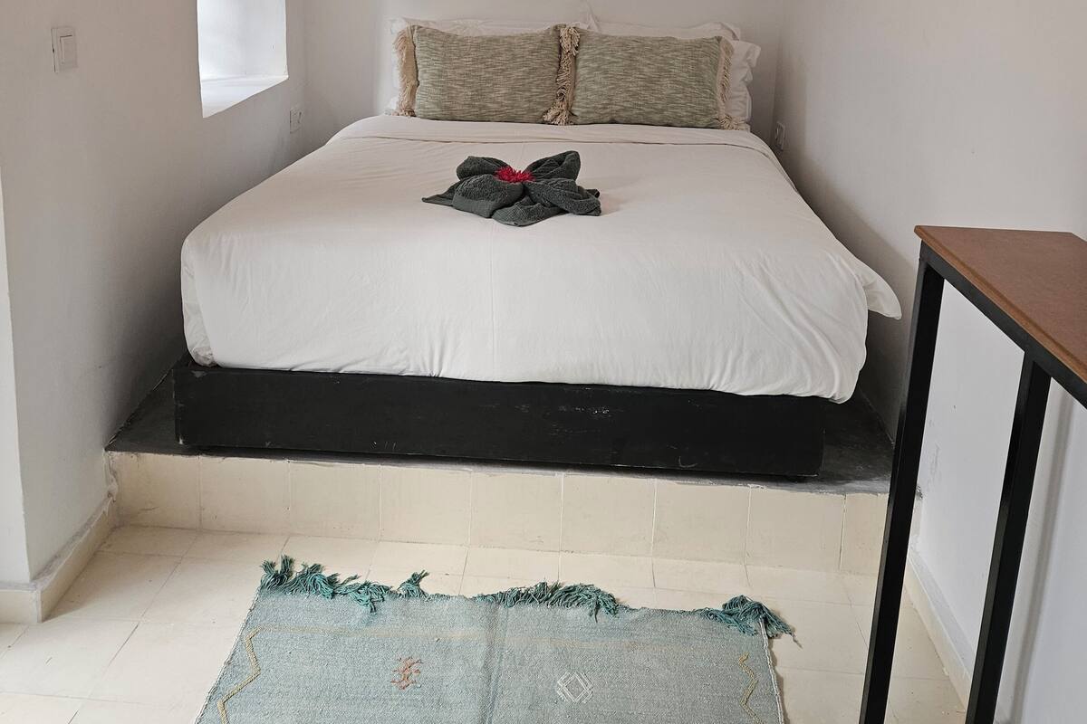 A bedroom with a bed, a window, a woven wall hanging, and a small table.