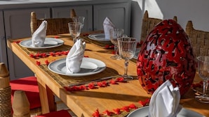 Taula de dinar posta amb plats blancs, servilletes i gots de vi transparents, envoltada de cadires de vimet i un bol decoratiu vermell.