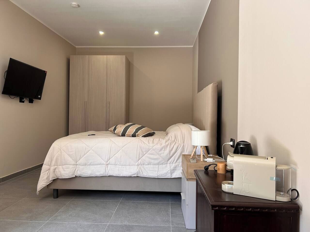 A modern hotel room with a bed, bedside table, lamp, coffee maker, and a flat-screen TV mounted on the wall.