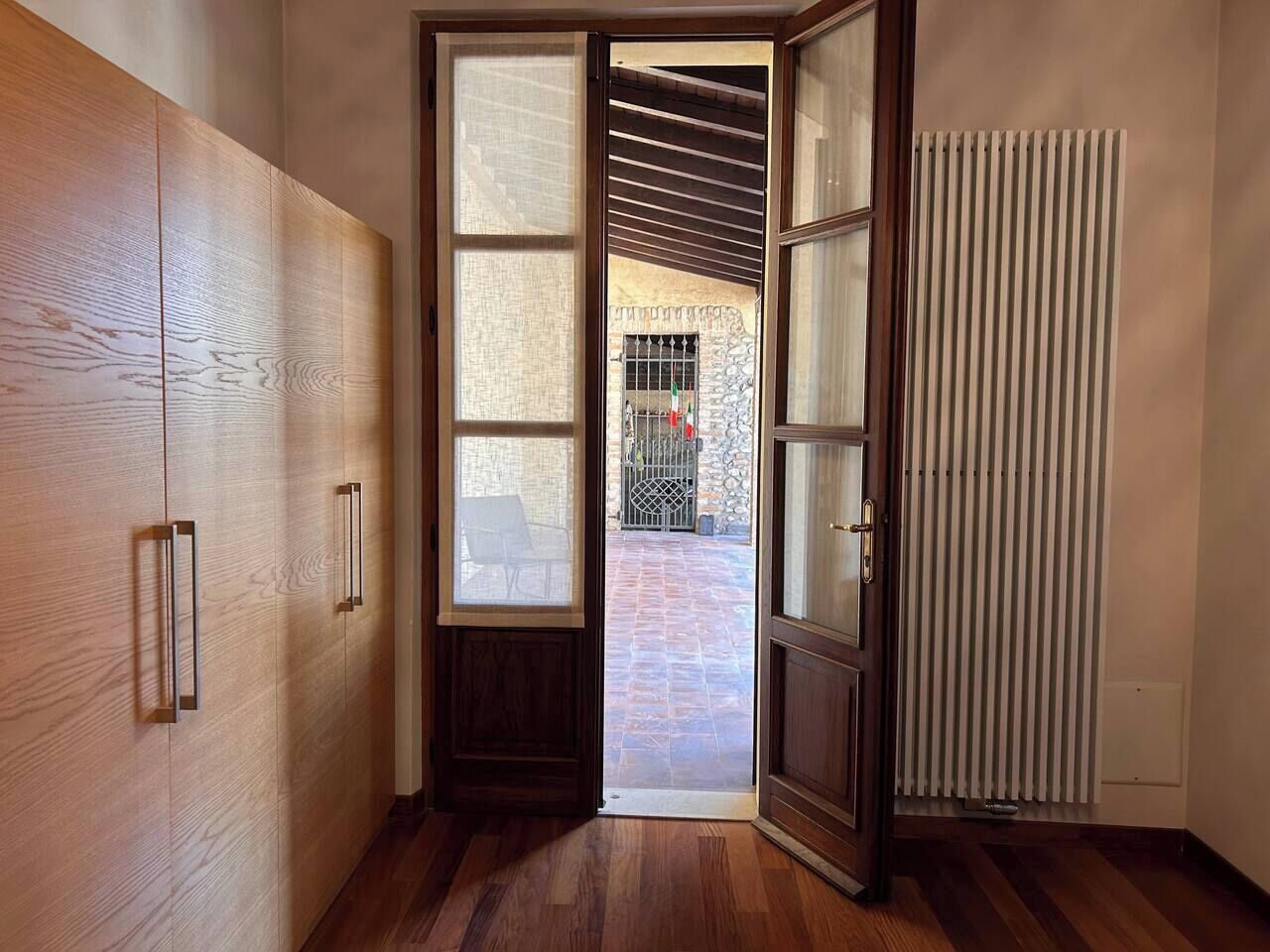 Un couloir doté d’un plancher en bois, une porte-fenêtre coulissante donnant sur un patio extérieur avec un mur en pierre et un portail en métal, et un radiateur sur le côté droit.