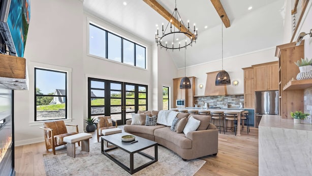 A spacious living room with a large sofa, a coffee table, and a fireplace. There is a kitchen area with wooden cabinets and a large island. The room has large windows and a chandelier.
