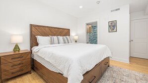 Un dormitorio con cabecera de madera, cubrecama blanco, dos mesitas de noche con lámparas, cómoda de madera y una puerta que da a otra habitación.