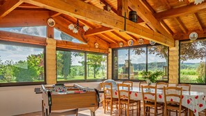 A wooden dining area with a table and chairs, large windows with a view of greenery, and a ceiling with exposed beams.