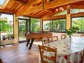A wooden-floored room with a pool table, a dining table with chairs, and large windows offering a view of greenery.
