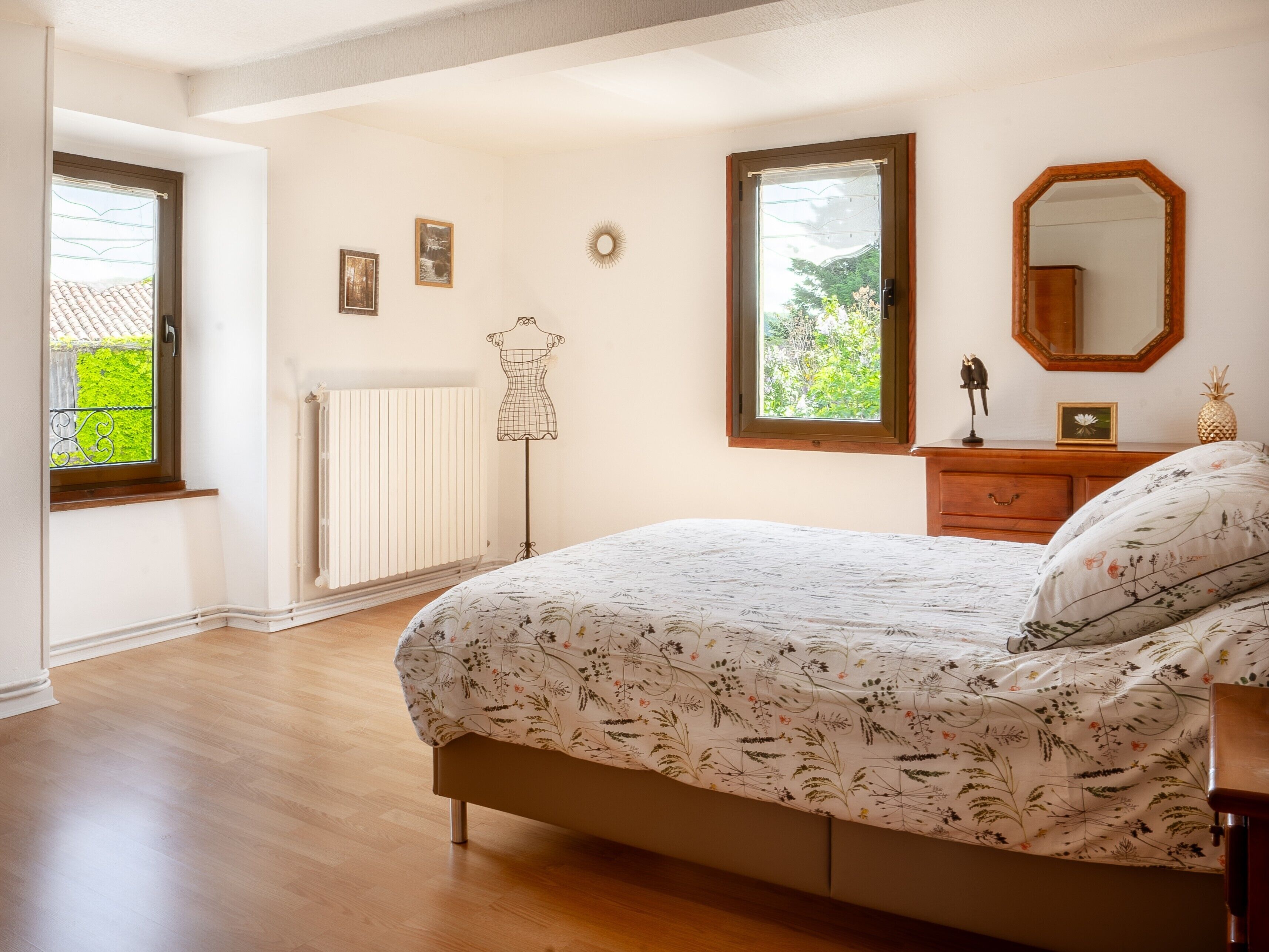 Een slaapkamer met een bed, een dressoir, een spiegel, een radiator en een raam met uitzicht op groen.
