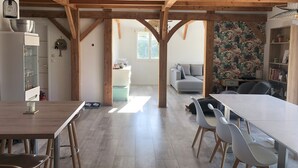 Eine geräumige Küche mit Holzbalken, einem Essbereich mit einem langen Tisch und Stühlen, einem grauen Sofa und einem Bücherregal.