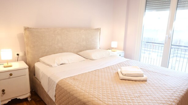 A neatly made bed with a quilted cover, two bedside lamps, a white nightstand, and a window with blinds.