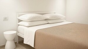 Una cama bien tendida con ropa blanca y un plaid de cuadros, una mesita de noche blanca y una pared lisa con un interruptor de luz.