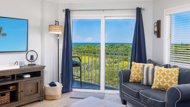 Un balcon avec vue sur l’océan, un canapé agrémenté de coussins à motifs, une lampe et un téléviseur posé sur un meuble en bois.