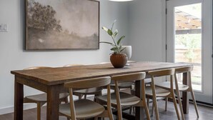 Un comedor con una mesa de madera, seis sillas, una planta en maceta, una lámpara colgante y un cuadro abstracto de gran tamaño en la pared.