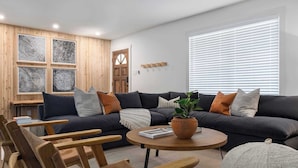 Una sala moderna con un sofá oscuro, sillas de madera, una mesa de centro redonda y un ventanal con persianas.