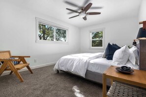 A bedroom with a bed, two windows, a ceiling fan, a wooden chair, and a small table.
