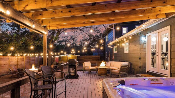 Una terraza al aire libre con jacuzzi, sillas de madera, una parrilla y luces colgantes.