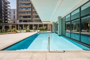 A modern indoor swimming pool with clear glass walls, surrounded by a tiled patio and outdoor seating.