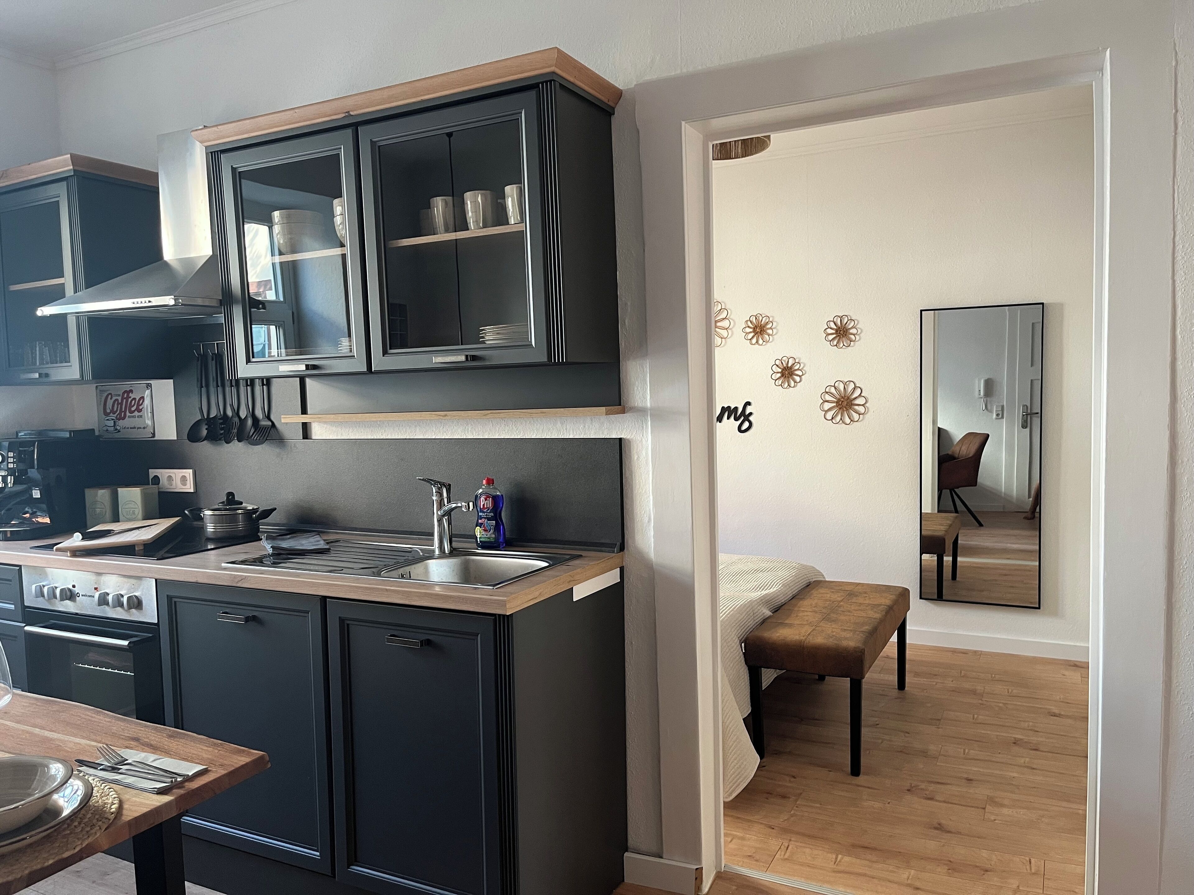 A compact kitchen with dark cabinets, a sink, and a stove. A dining table with cutlery is visible, and a mirror reflects part of the room.