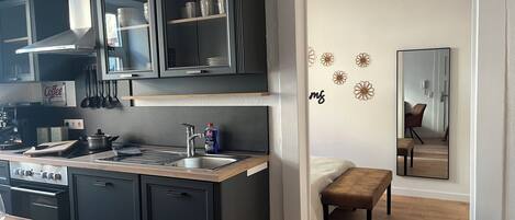 A compact kitchen with dark cabinets, a sink, and a stove. A dining table with cutlery is visible, and a mirror reflects part of the room.