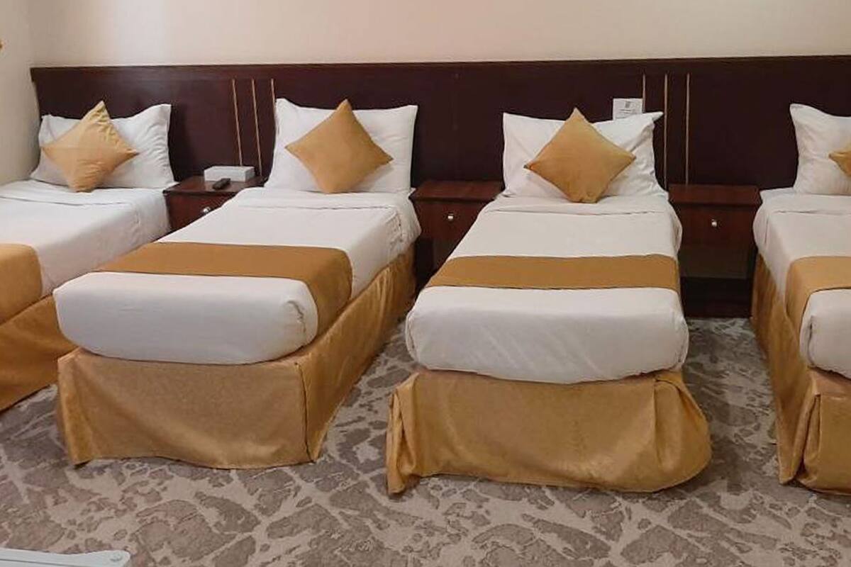 A hotel room with four single beds, each with a brown bed skirt and white bedding. The room has a patterned carpet and a wooden headboard.