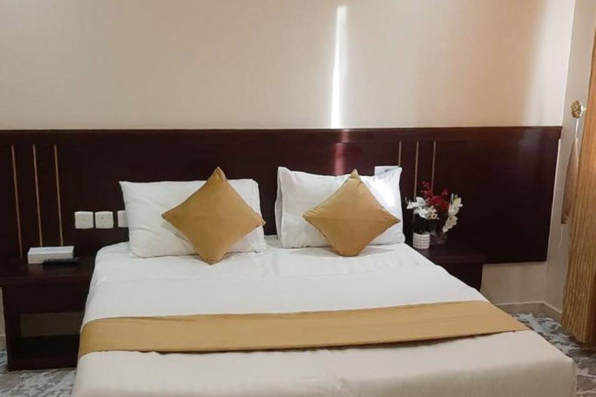 A hotel room with a large bed, two white pillows, a gold throw blanket, and a wooden headboard.