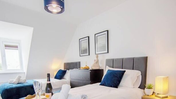 A modern bedroom with a large bed, a smaller bed, a bedside table with a lamp, and a ceiling light fixture.