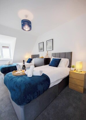 Ein modernes Schlafzimmer mit einem großen Bett, einem kleineren Bett, einem Nachttisch mit Lampe und einer Deckenleuchte.