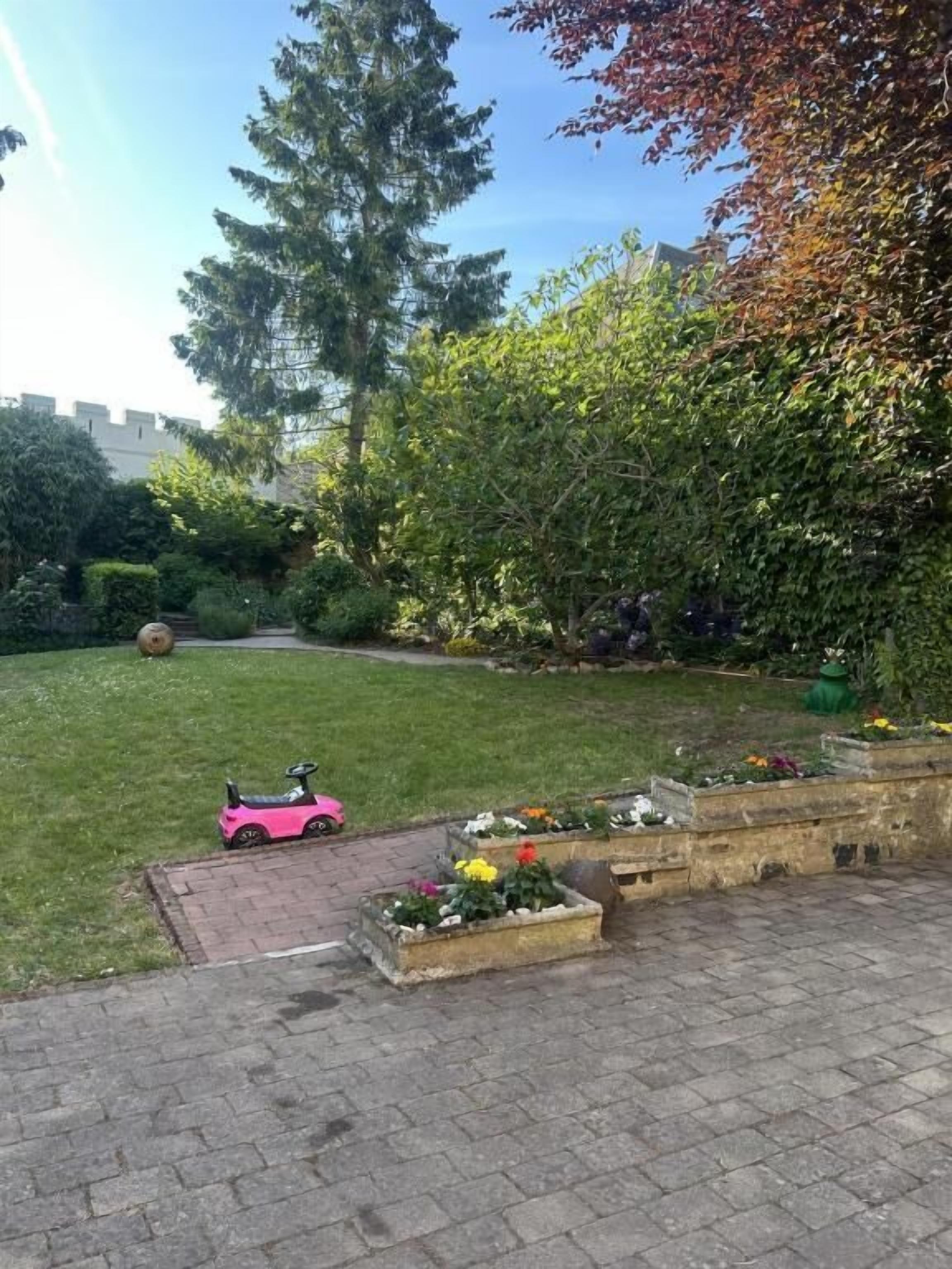 A paved patio area with a pink toy car, a grassy lawn, and a variety of plants and trees.