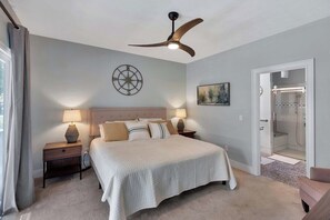 Quarto com cama grande, luminárias de cabeceira, ventoinha no teto, relógio de parede e banheiro.