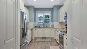 Una cocina con armarios blancos, un refrigerador de acero inoxidable, un lavamanos doble y una ventana con vistas a árboles.