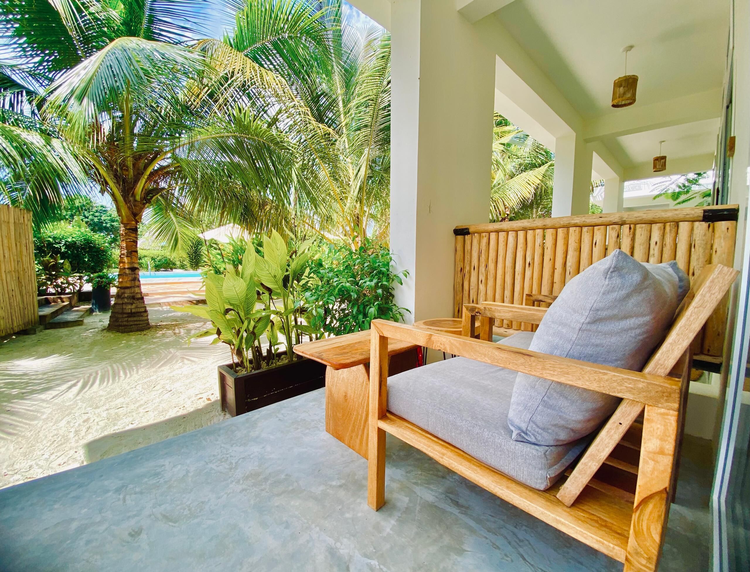 A wooden bench with a cushion in a covered outdoor area with a palm tree and potted plants.