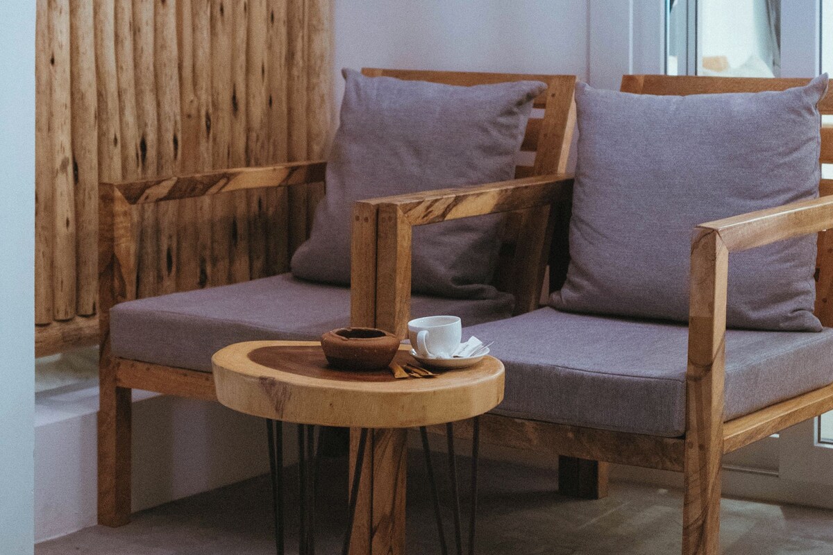 Un sofá de madera con cojines, una mesita redonda pequeña con una taza y platillo, y un piso de concreto.