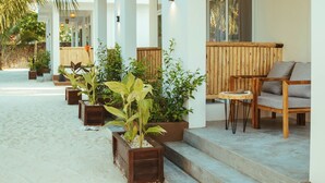 Een hotel aan het strand met terras, houten plantenbakken met groen en een zandpad.