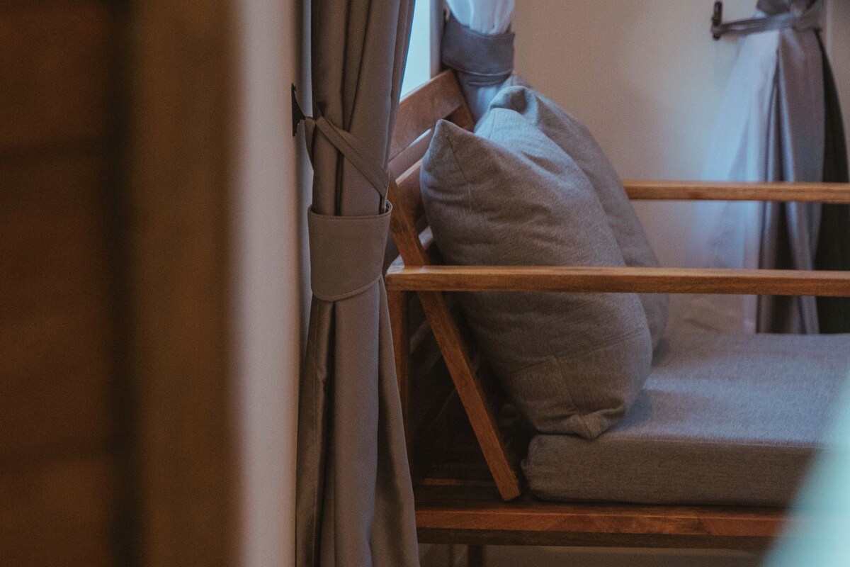 A room with a wooden bed, grey bedding, and curtains tied back.