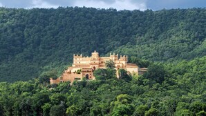 A large, historic building with multiple levels and a complex roof structure, surrounded by dense green forest.