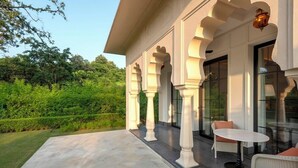 A covered outdoor area with columns, a table, and chairs, surrounded by greenery.