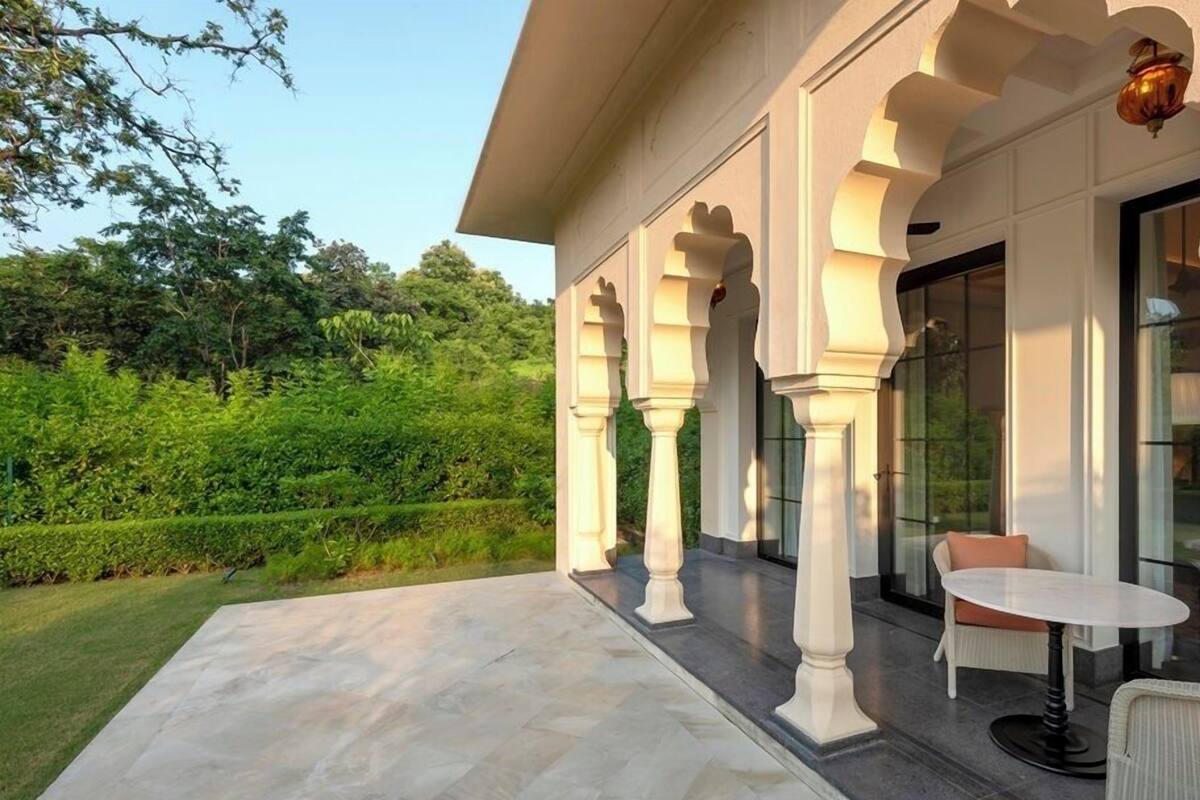 A covered outdoor area with columns, a table, and chairs, surrounded by greenery.