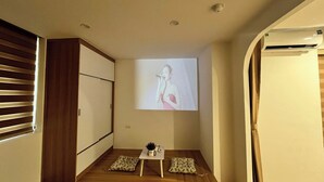 Una habitación con suelo de madera, una pequeña mesa con una planta y una pantalla de proyección mostrando a una persona.