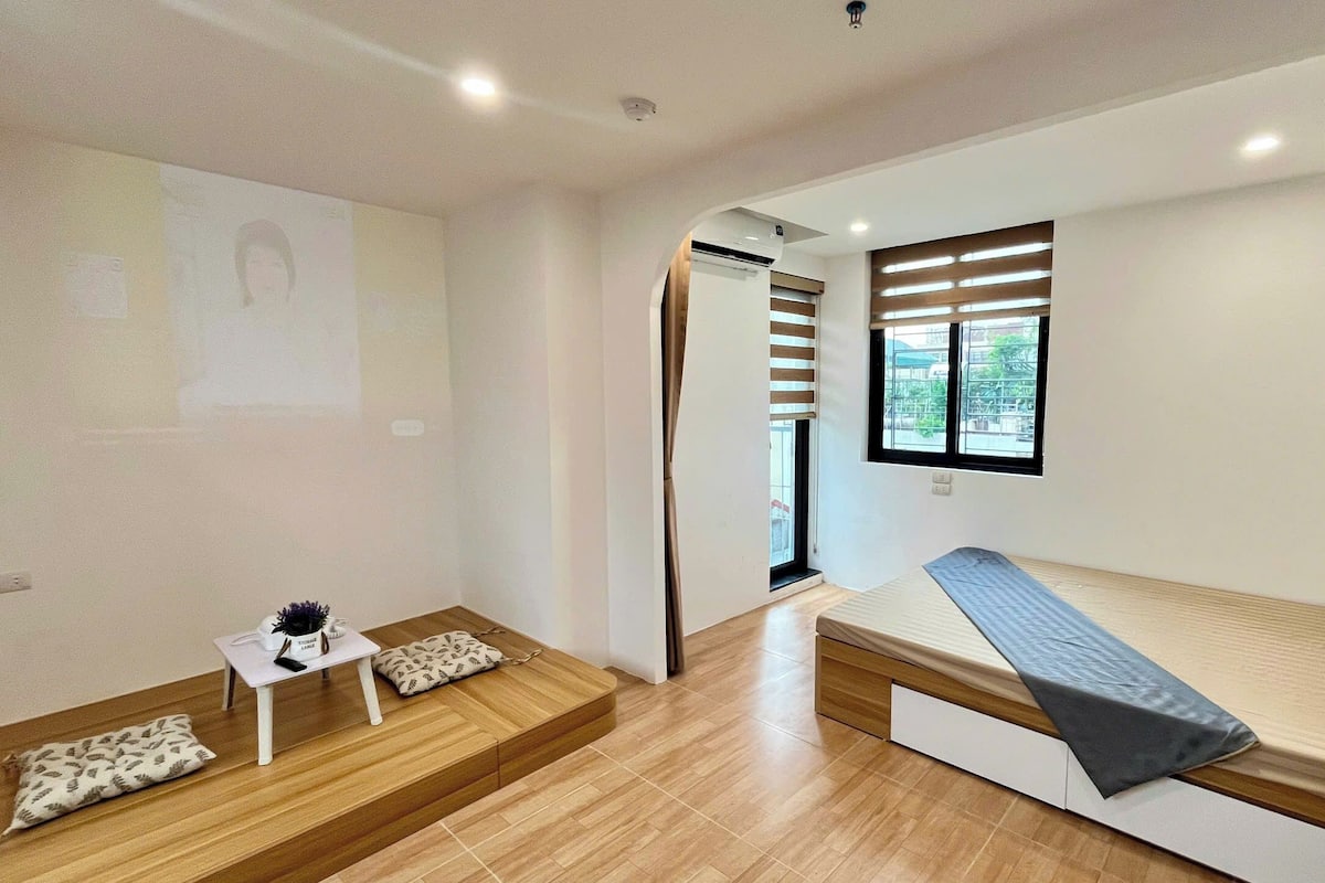 A compact room with a bed, a small table with a lamp, a wall-mounted air conditioner, and a window with blinds.
