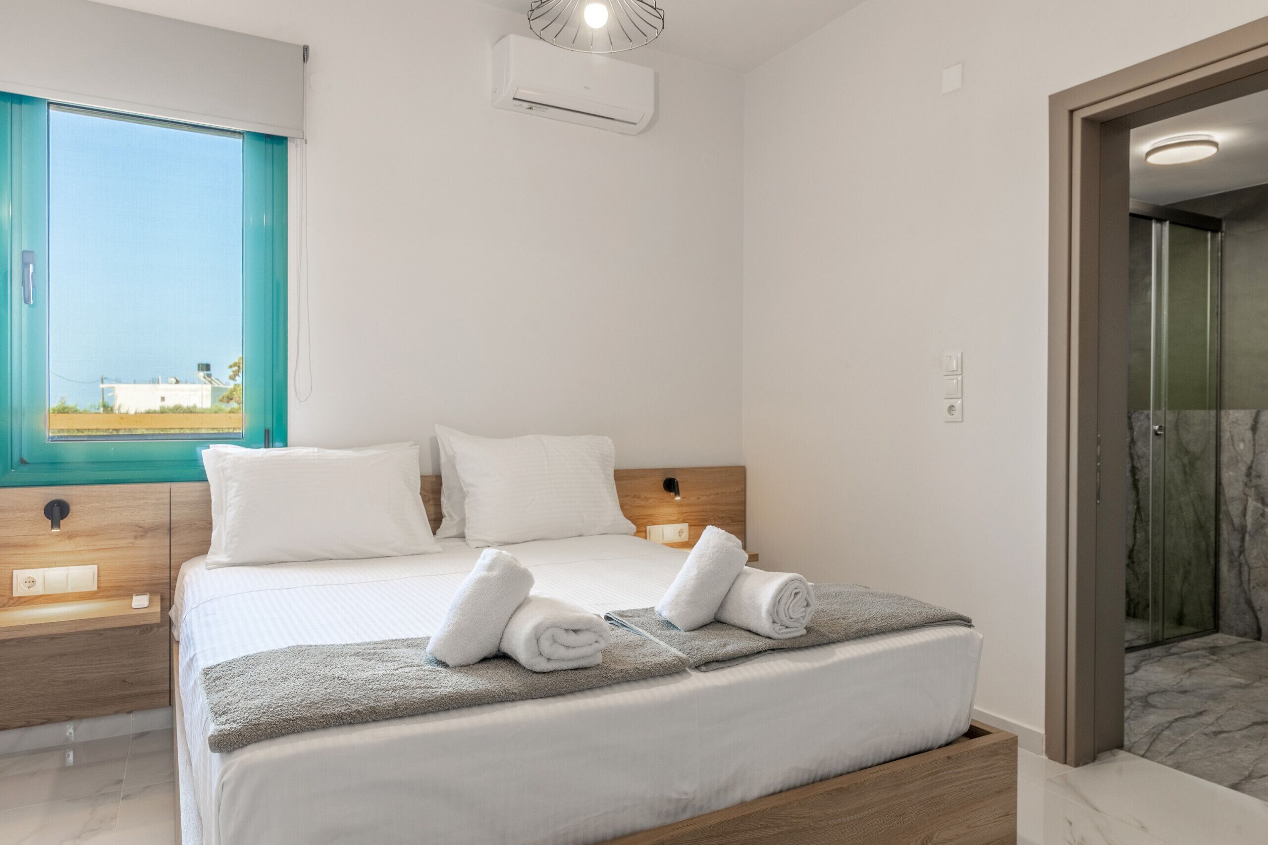 A modern hotel room with a bed, a wooden headboard, a window with a view, and a shower area.