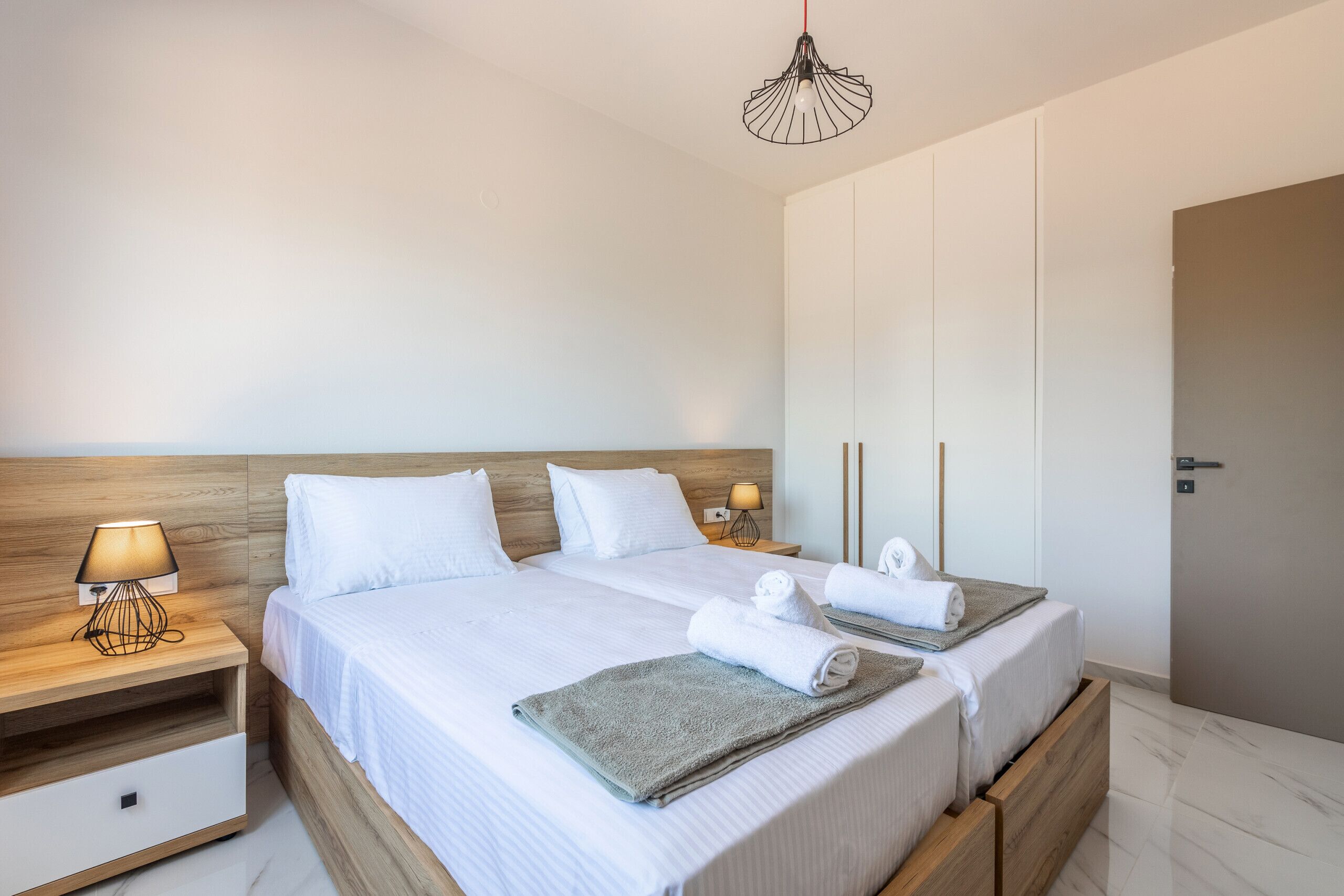 Cama doble con sábanas blancas, dos mesitas de noche con lámparas, cabecera de madera, manta gris y lámpara colgante.