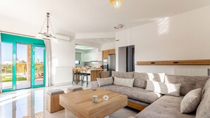 A modern living room with a sectional sofa, a wooden coffee table, and a view of the outdoors through large sliding glass doors. - Sandi Villa (Hersonissos)
